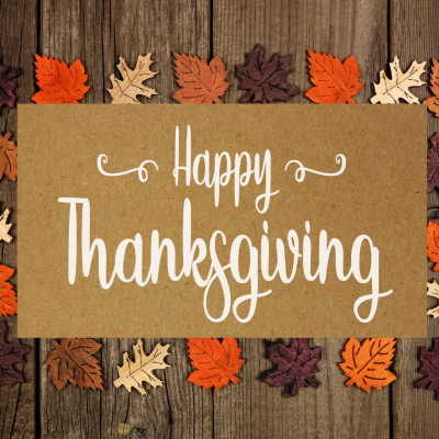A "Happy Thanksgiving" sign on a brown background, surrounded by variously colored autumn leaves on a wooden surface.