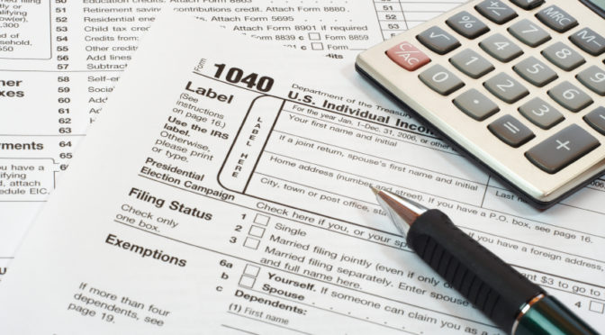 A pen and calculator rest on a stack of IRS 1040 tax forms and other documents, indicating tax preparation work.