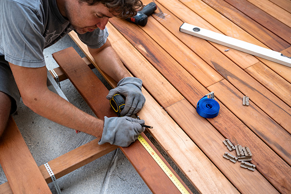 A person wearing gloves measures and marks wooden planks with a tape measure on a decking project, with tools and fasteners nearby.