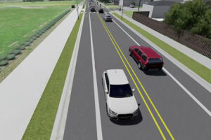 A computer-generated image shows several cars driving on a two-lane road with a double yellow line, bordered by sidewalks, grass, and residential buildings.