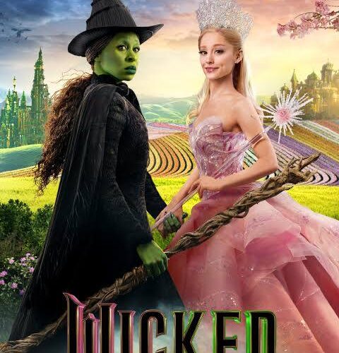 Two women in costume stand side by side, one dressed as a green-skinned witch in black, the other in a pink gown and crown, with a colorful fantasy landscape in the background. 