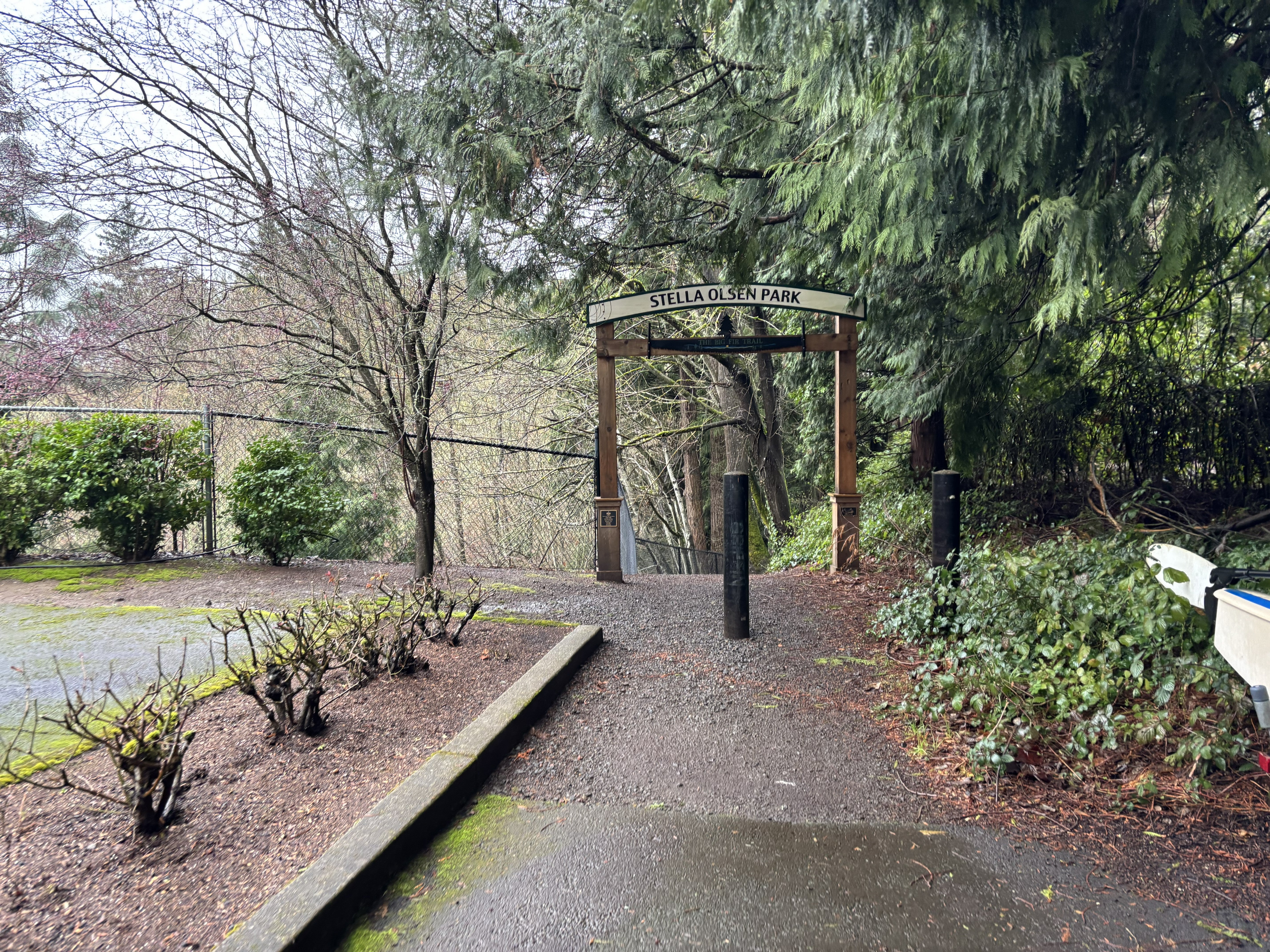 A wooden entrance sign for Stella Olsen Park stands among trees and bushes along a wet, paved path.