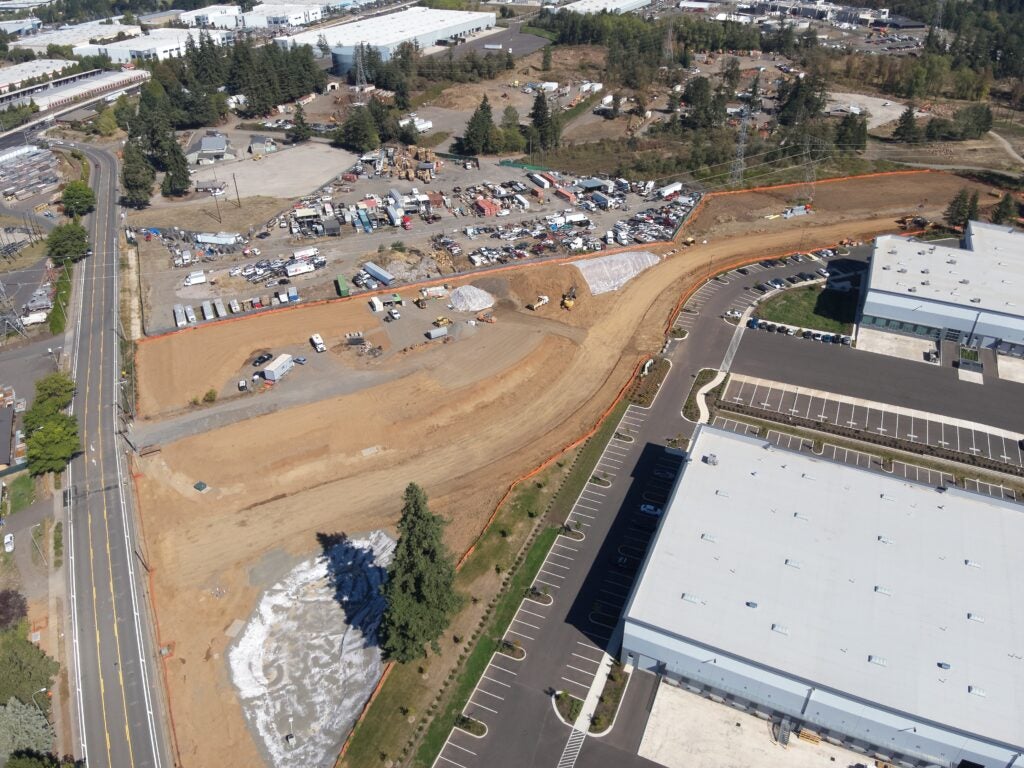 Aerial view of a construction site with cleared land, construction equipment, and materials next to a large warehouse and parking lot.