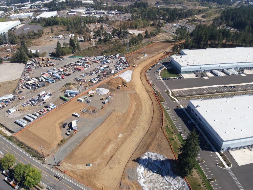 Aerial view of a construction site with heavy machinery, adjacent to parking lots, warehouses, and a nearby street, surrounded by trees and industrial buildings.