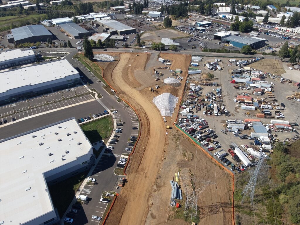 Aerial view of a construction site with grading equipment, dirt roadwork, nearby industrial buildings, parked vehicles, and utility towers.