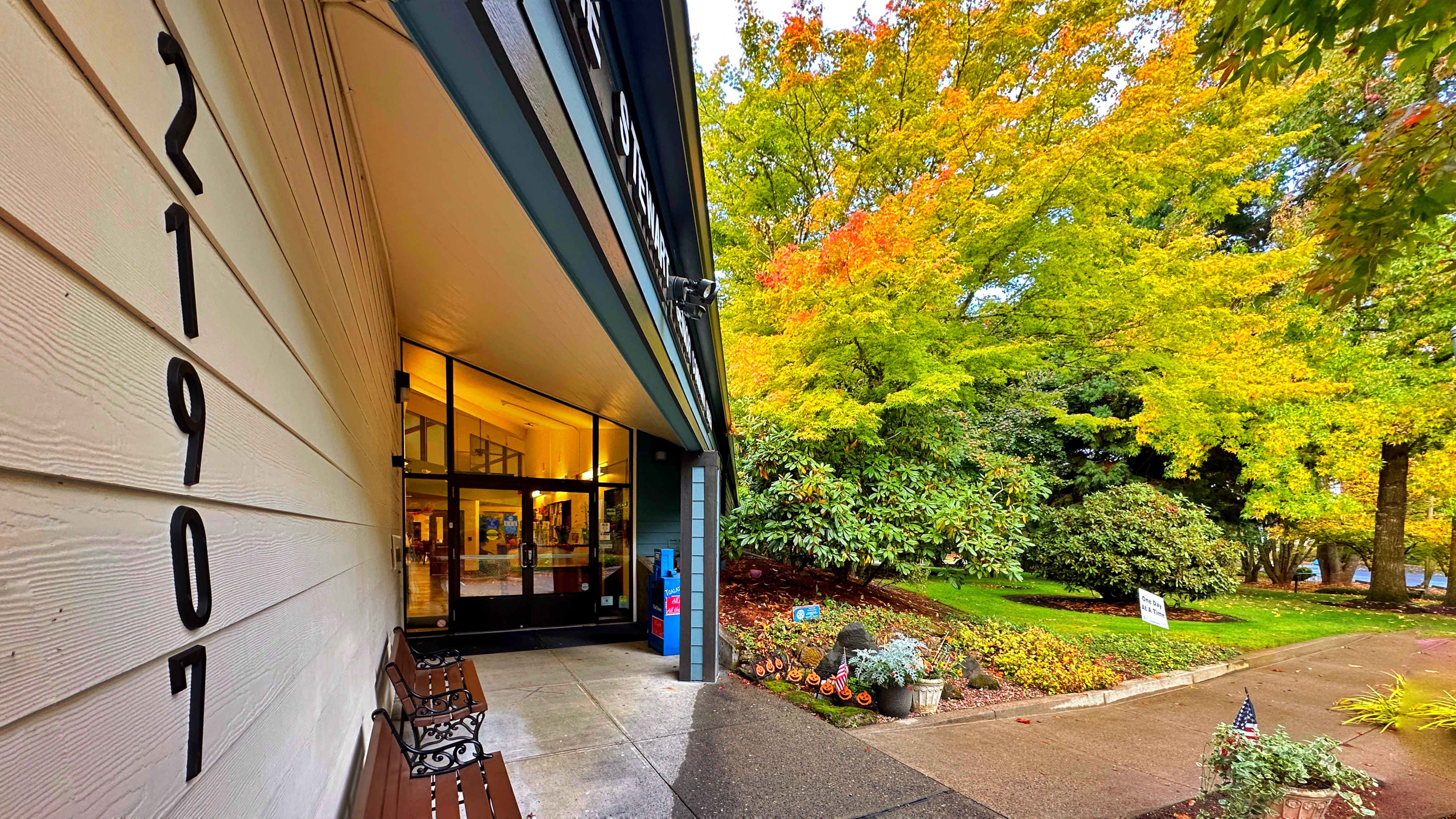 Building entrance with large glass doors, address 21907 on the wall, a bench, and vibrant autumn trees in a landscaped area.