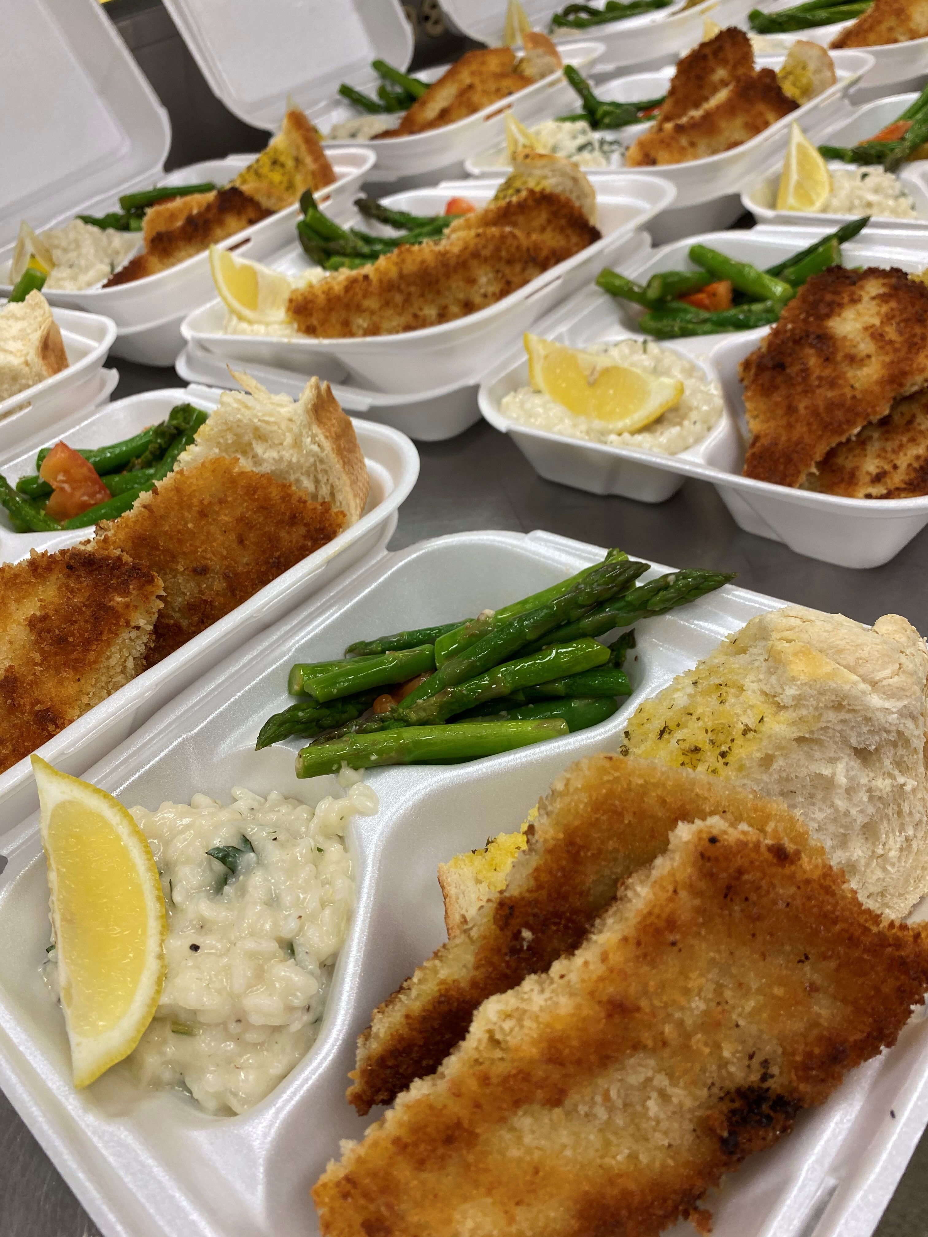 This is a photo of several to-go meals in  three section Styrofoam delivery containers. In one section is a lemon wedge on top of risotto, the right section holds fresh cooked green means, and the main section holds two hand-breaded pieces of cod.