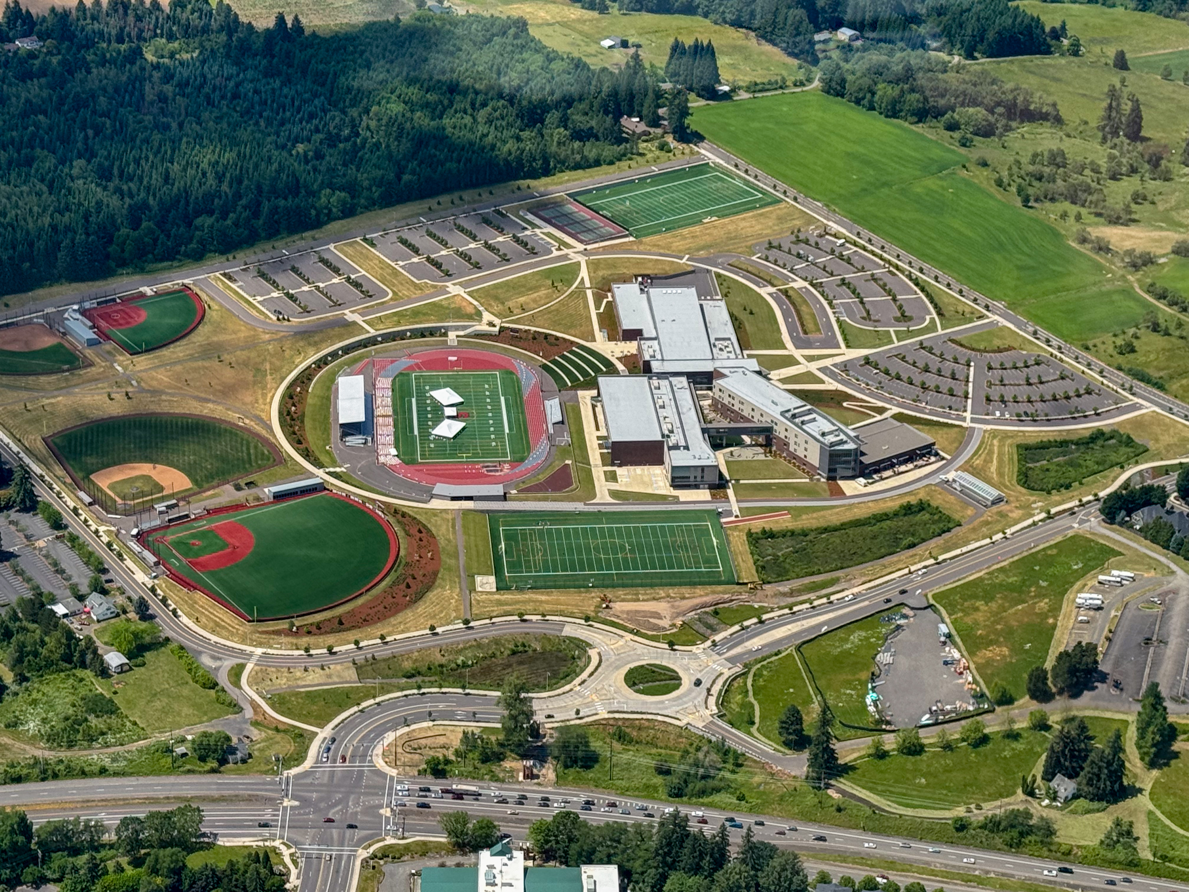 Aerial view of a large school campus with multiple sports fields, a track, parking lots, circular building layout, and surrounded by green landscape.