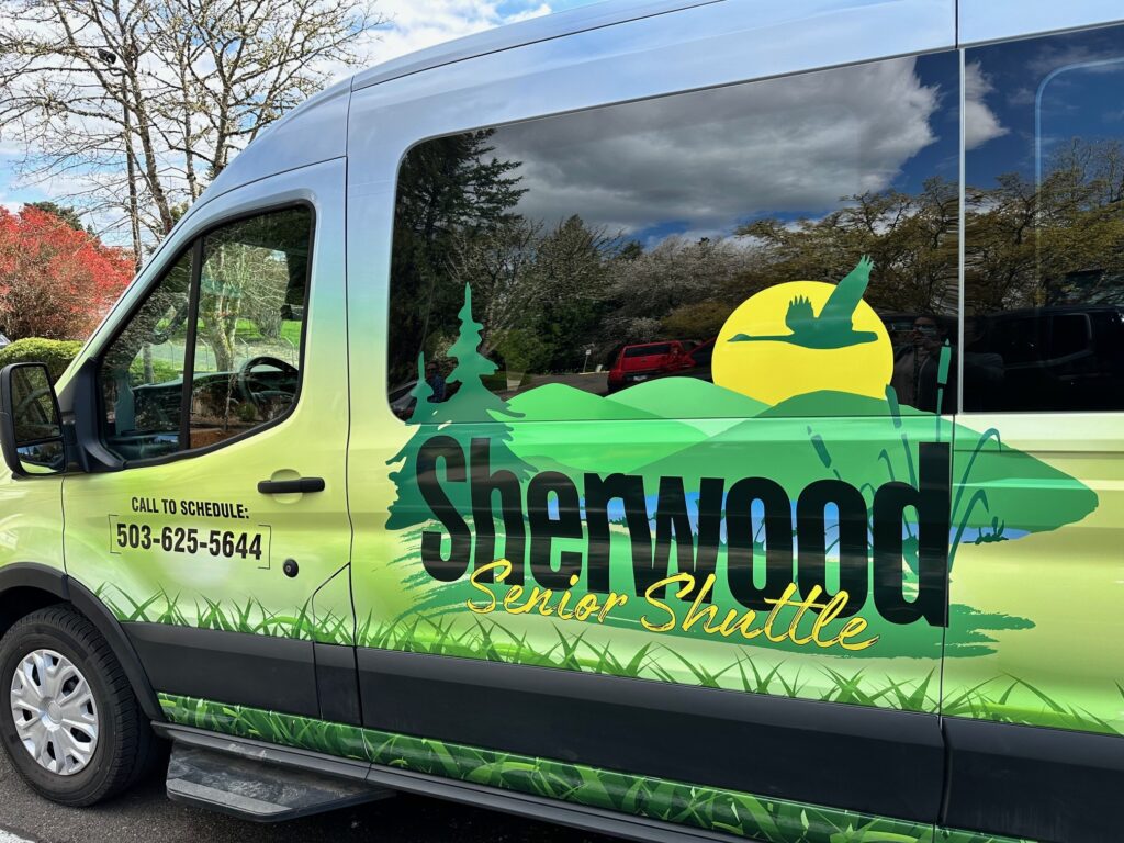 This photo depicts the Sherwood Senior Shuttle, a van that has been painted with a blue sky and light green fields. On the side of the van is the Sherwood Shuttle logo featuring three medium green pine treen on the right, three light green hills, and a yellow sun with the silhouette of a flying Canadian goose. 