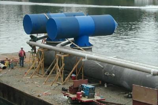 Large blue cylindrical structures with attached pipes are set up near the water’s edge; a person stands nearby for scale.