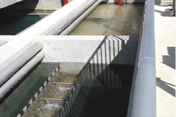Concrete water treatment channel with metal grates and pipes, carrying water through a filtration or separation process in an industrial facility.