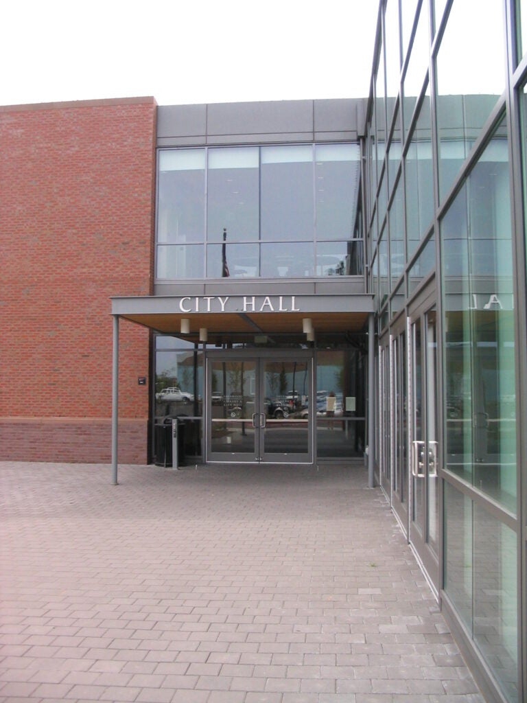 Glass entrance to a modern city hall building with "City Hall" sign above the doors, adjacent to a brick wall and paved walkway.