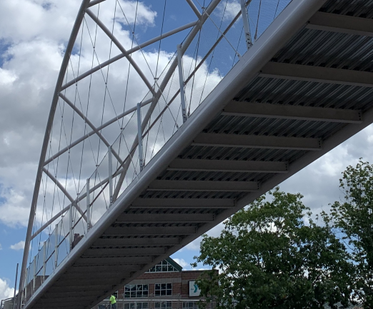 A modern pedestrian bridge with metal framework and cables spans overhead, with a partly cloudy sky and trees visible in the background.