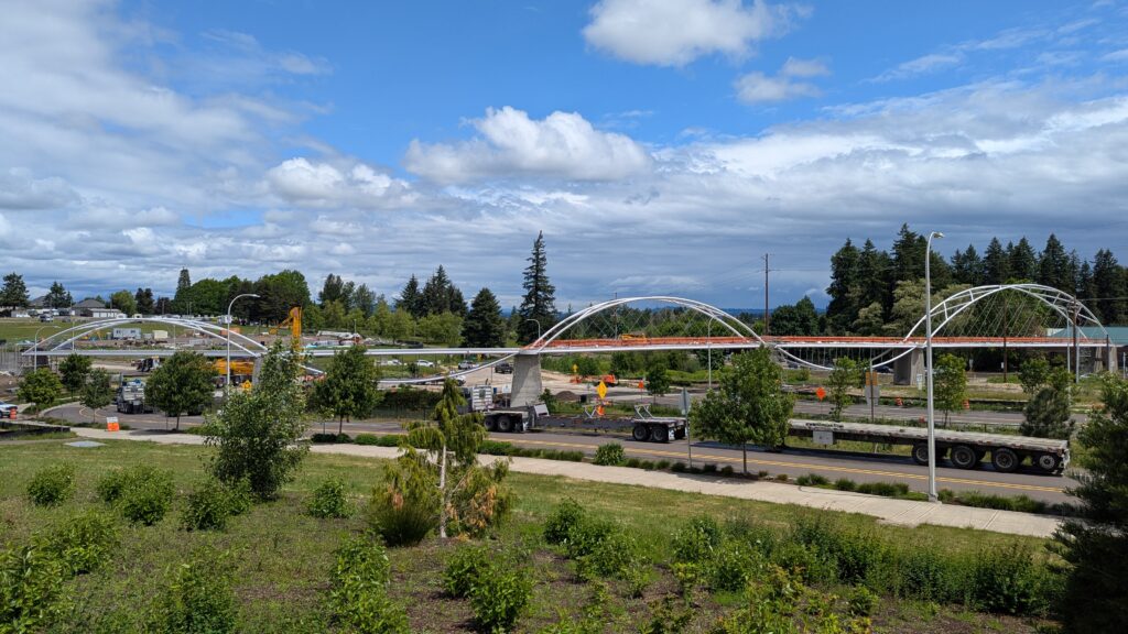 Pedestrian bridge under construction spans a road, with cranes, trucks, and construction equipment present amid a landscaped, green area under a partly cloudy sky.