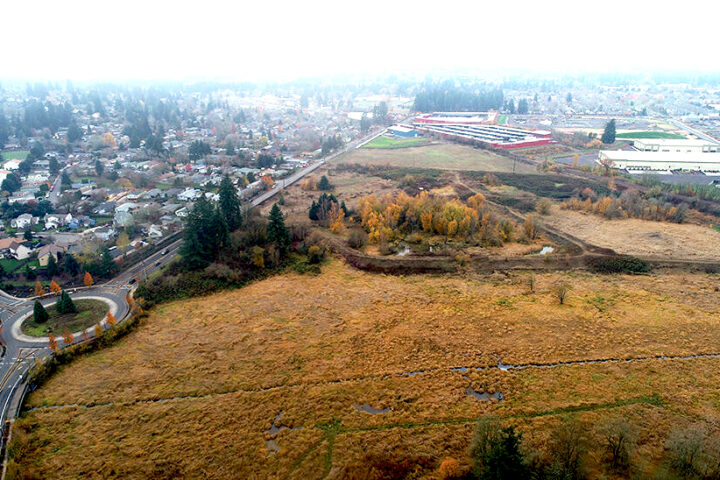 Aerial view of a grassy, open field bordered by trees, with a roundabout and residential neighborhood on the left, and buildings visible in the distance.