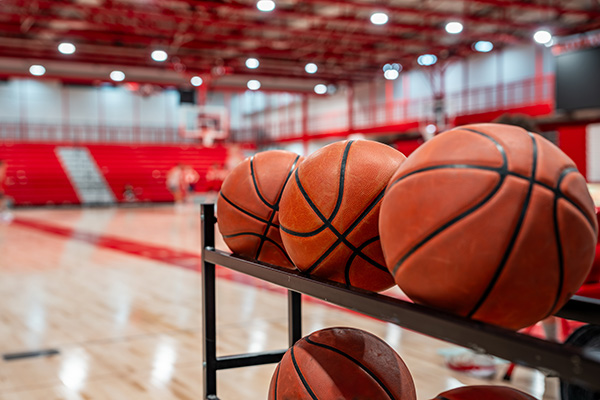 Several basketballs are lined up on a rack in a gymnasium with a basketball court and red bleachers in the background.
