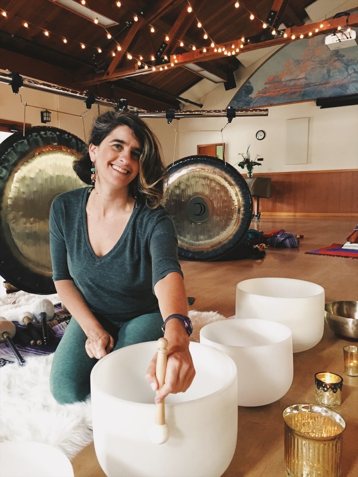 A woman sits on the floor indoors, smiling, and playing a white crystal singing bowl with a mallet; gongs and candles are visible in the background.