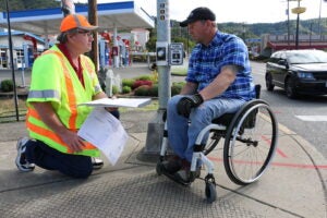 A construction worker and a man in a wheelchair discuss plans at a street corner by a crosswalk button, with buildings and cars in the background.