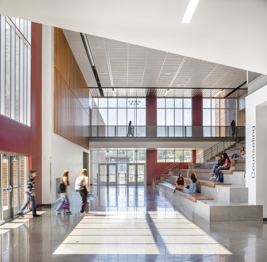 A modern school lobby with large windows, concrete seating steps, several people walking, and some students sitting and talking near a counseling sign.