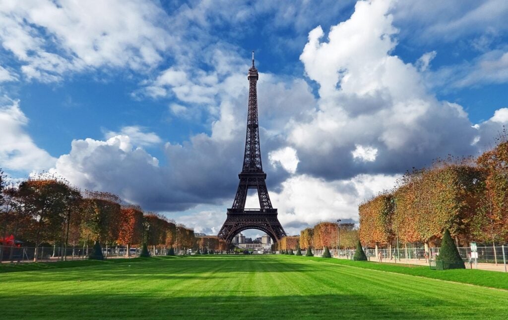 The Eiffel Tower stands centered under a partly cloudy sky, bordered by trimmed trees and a large green lawn in the foreground.