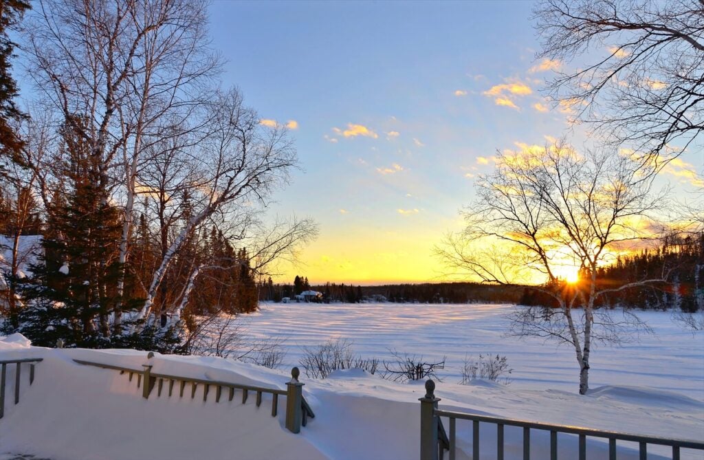 Sun sets over a snowy, frozen landscape surrounded by leafless trees and evergreens, viewed from behind a snow-covered wooden railing.