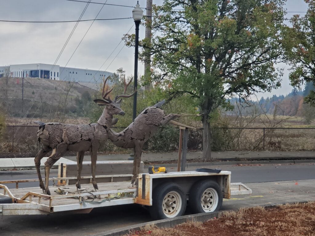 Two metal deer sculptures are displayed on a trailer parked by a roadside, with trees and an industrial building visible in the background.