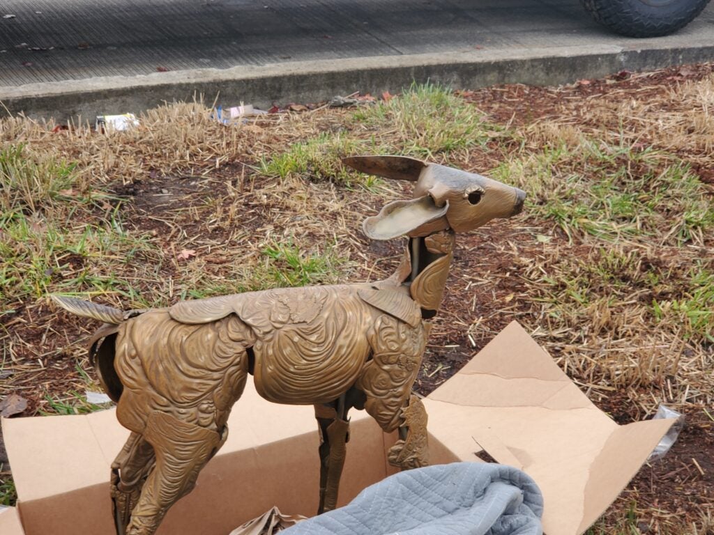 A metal deer sculpture stands in a cardboard box on the ground near a patch of grass and a sidewalk.