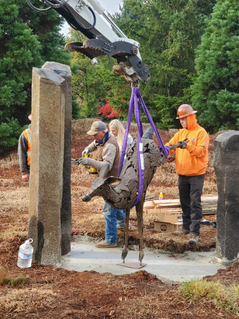 Workers install a metal animal sculpture using a crane and straps between two stone pillars in an outdoor setting.