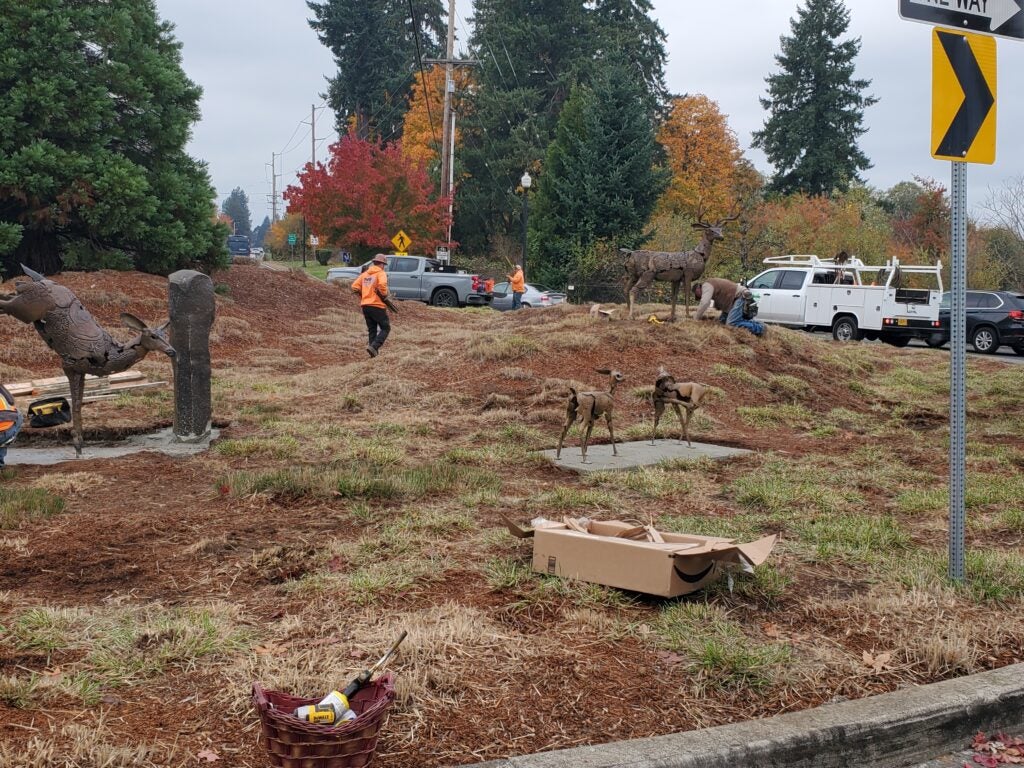 Workers in orange vests install metal deer sculptures on a grassy roadside as trucks and autumn trees are visible in the background.