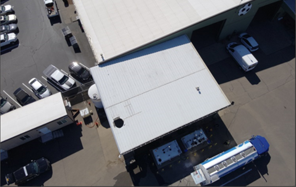 Aerial view of an industrial facility with several parked vehicles, a large white-roofed building, and equipment near the entrance.