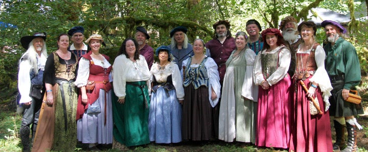 A group of people dressed in Renaissance-style costumes stand together outdoors in a wooded area, smiling at the camera.