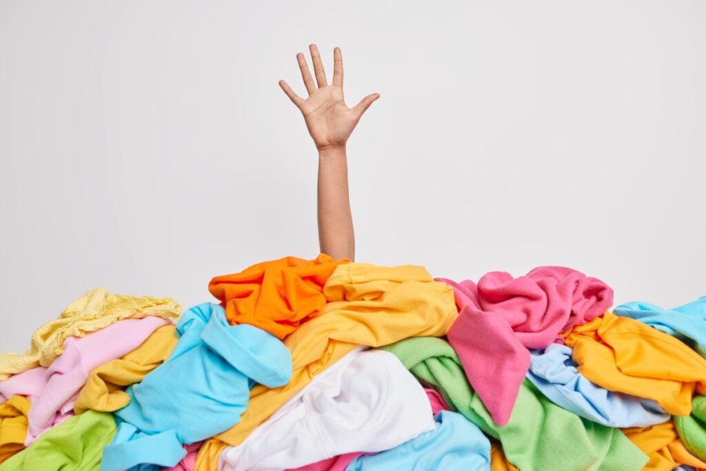 A hand reaches upward from a large pile of colorful laundry against a plain white background.