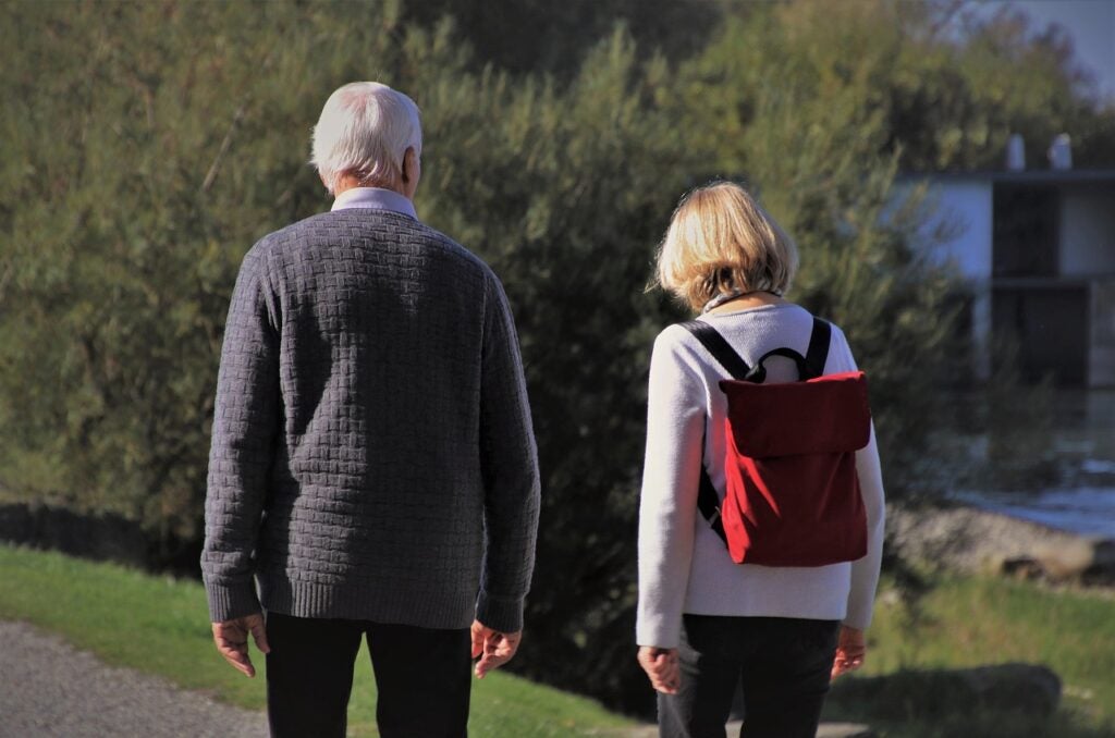 An older man and woman walk outdoors on a sunny day; the man wears a grey sweater and the woman has a red backpack and white sweater.