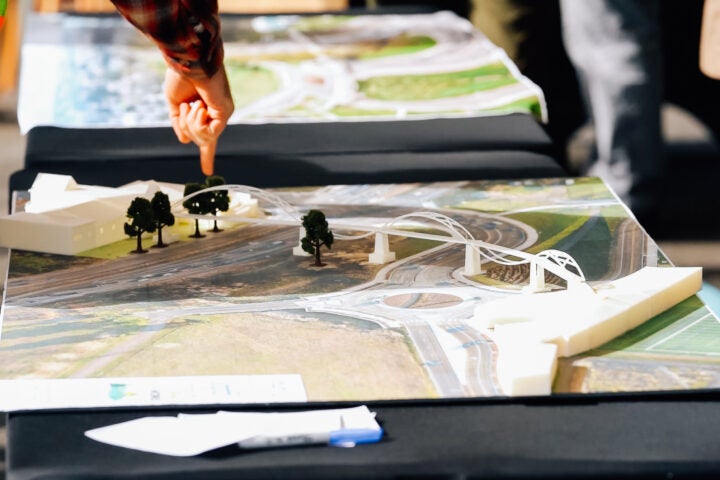 A person points to a section of a scaled architectural model showing roads, bridges, and trees on a table.