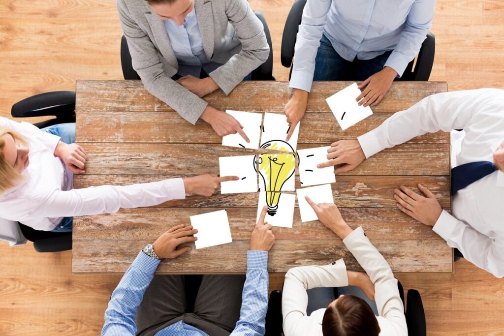 Six people sit around a wooden table, each holding a piece of paper forming a drawing of a light bulb in the center, symbolizing teamwork and collaboration.