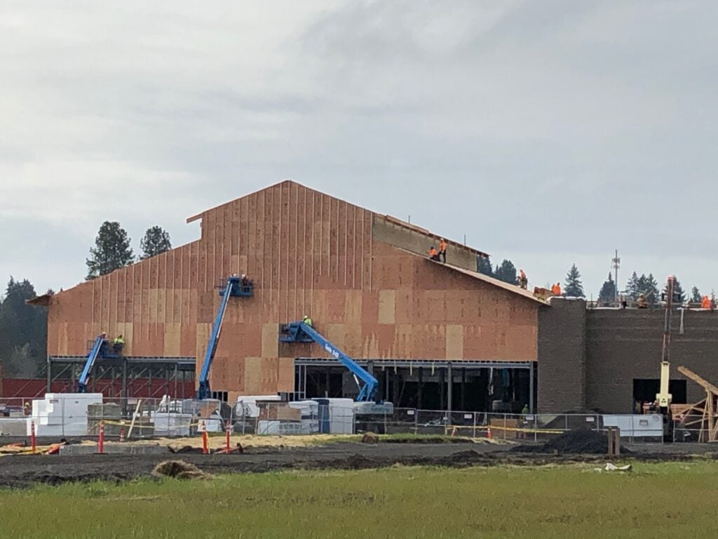 Construction site with workers on and around a large, partially finished building using lifts and scaffolding, with building materials and equipment in the foreground.