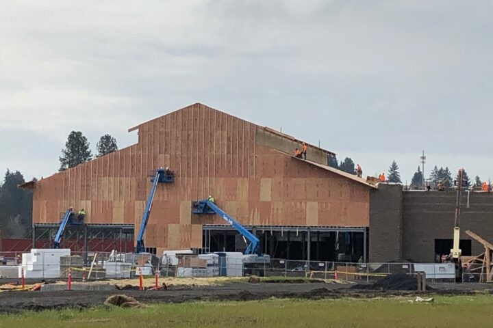 Construction site with workers on and around a large, partially finished building using lifts and scaffolding, with building materials and equipment in the foreground.