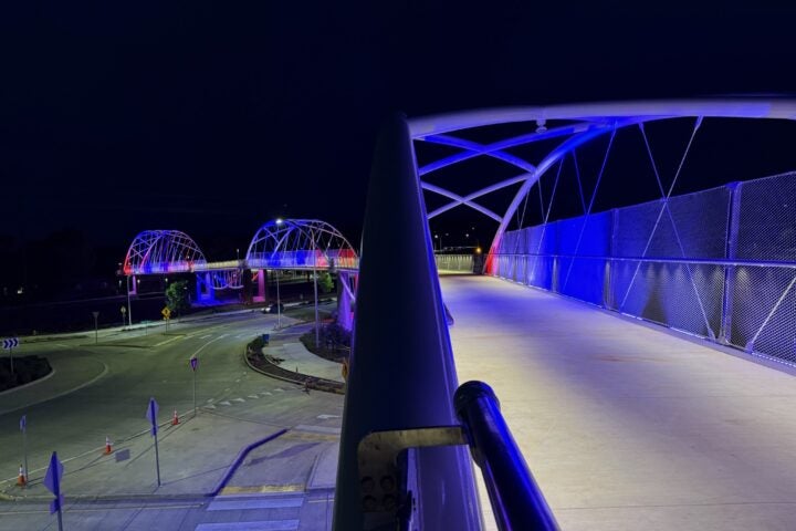A modern pedestrian bridge at night, illuminated with blue and purple lights, spans over a roundabout and connects to another lit bridge in the background.