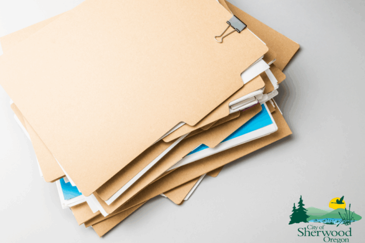 A stack of manila file folders is arranged on a white surface, with a binder clip on top. The City of Sherwood, Oregon logo appears in the bottom right corner.