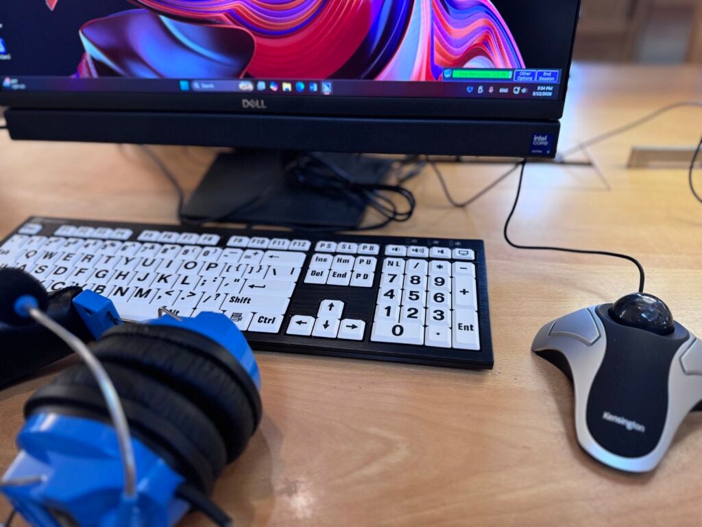 A desktop setup with a large print keyboard, a trackball mouse, blue headphones, and a Dell monitor displaying a colorful screen.