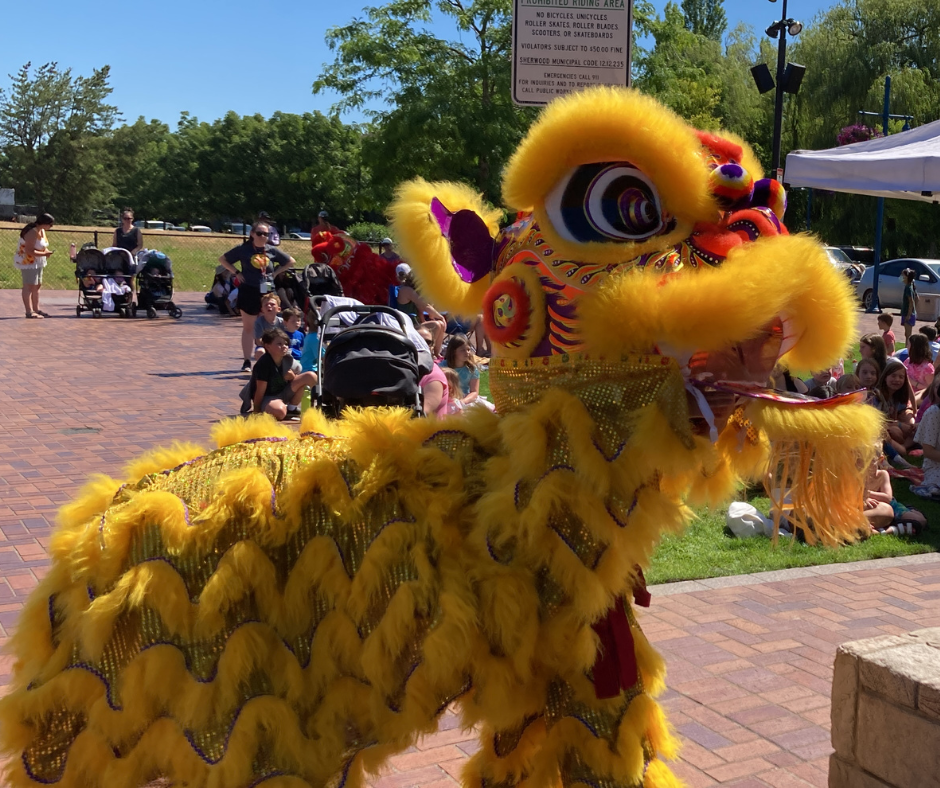 A yellow lion costume is used in a lion dance performance outdoors, with a crowd watching and strollers in the background.