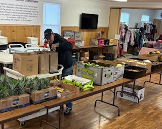 A person packs food items into bags on a table filled with fresh produce and groceries inside a community center with clothes hanging in the background.