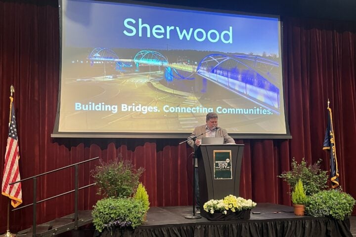 A person speaks at a podium on a stage with plants, in front of a screen displaying 