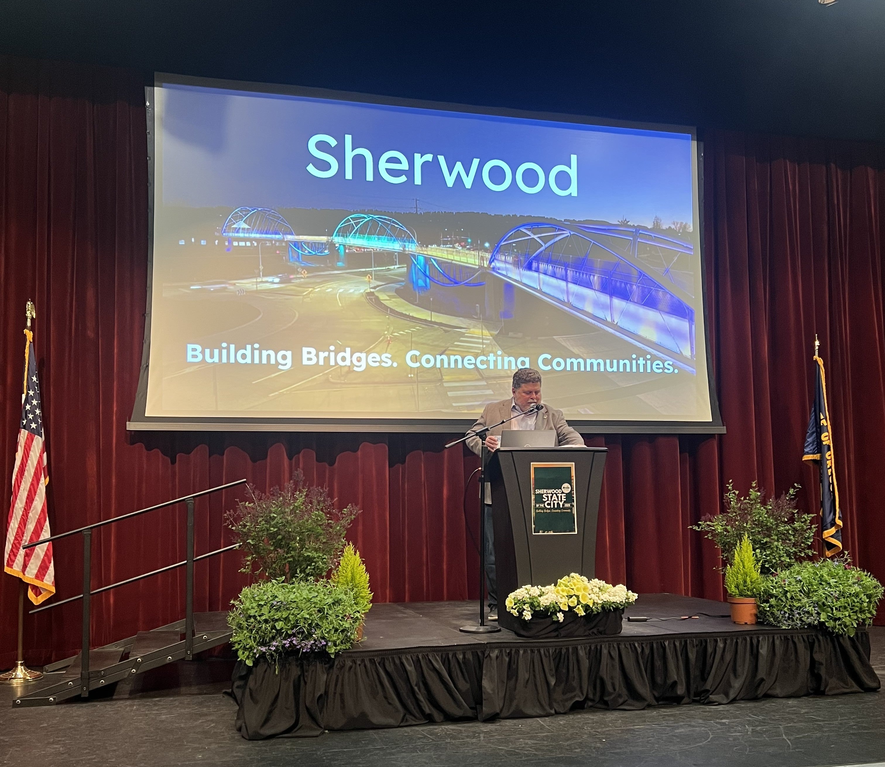 A person speaks at a podium on a stage with plants, in front of a screen displaying 