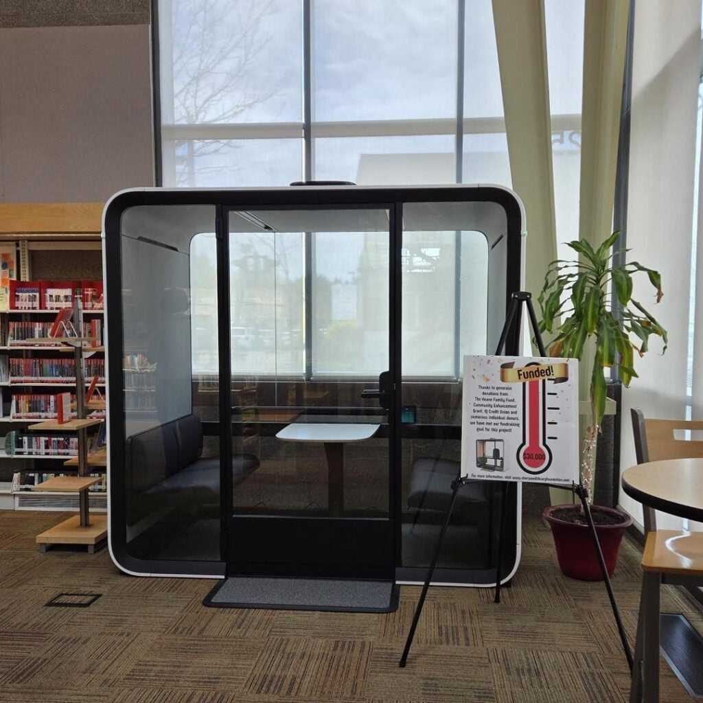 A soundproof meeting pod with a table and benches sits in a library near a window; a sign with a funding thermometer is displayed beside it.