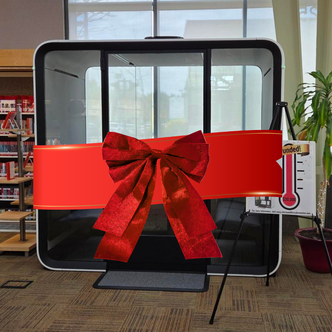 A soundproof booth in a library is decorated with a large red ribbon and bow, with a fundraising thermometer sign beside it.