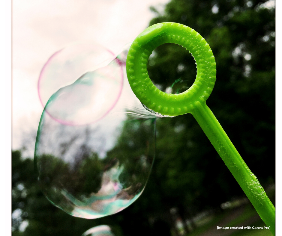 A green bubble wand is shown with a soap bubble forming in front of a blurred outdoor background with trees and sky.