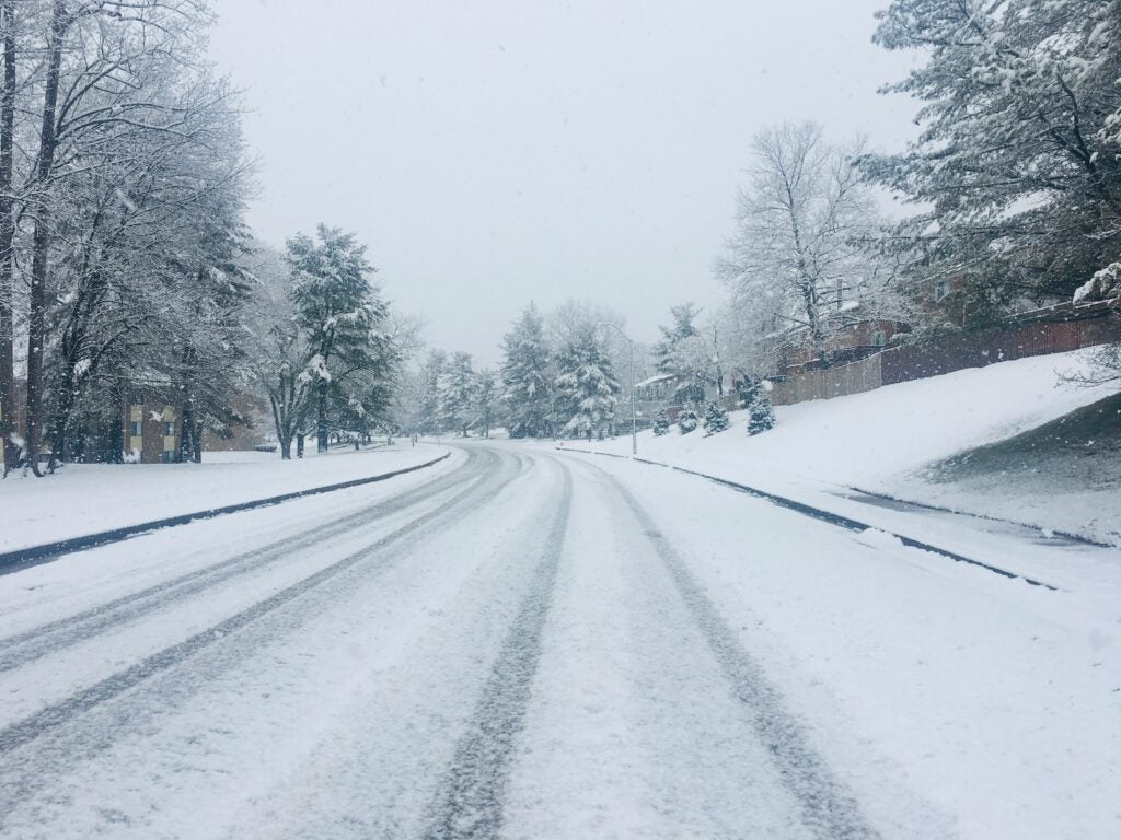 A snow-covered road curves through a suburban area with trees and houses lining both sides, on a cloudy winter day.