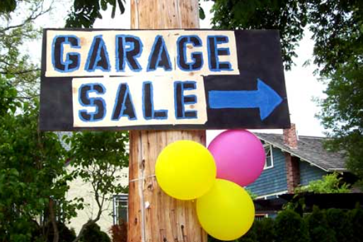 A garage sale sign with a blue arrow is attached to a wooden pole, decorated with yellow and pink balloons. Trees and a house are visible in the background.