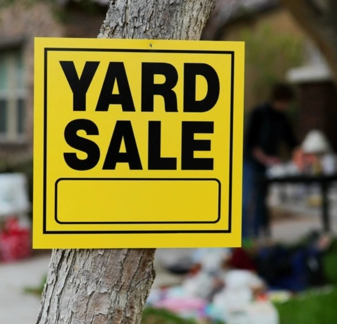 A yellow "Yard Sale" sign is posted on a tree, with items and a person visible in the blurred background.
